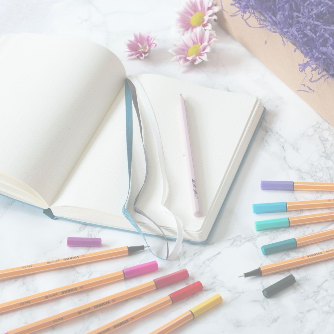 Colorful markers on a marble surface with an open notebook and flowers.