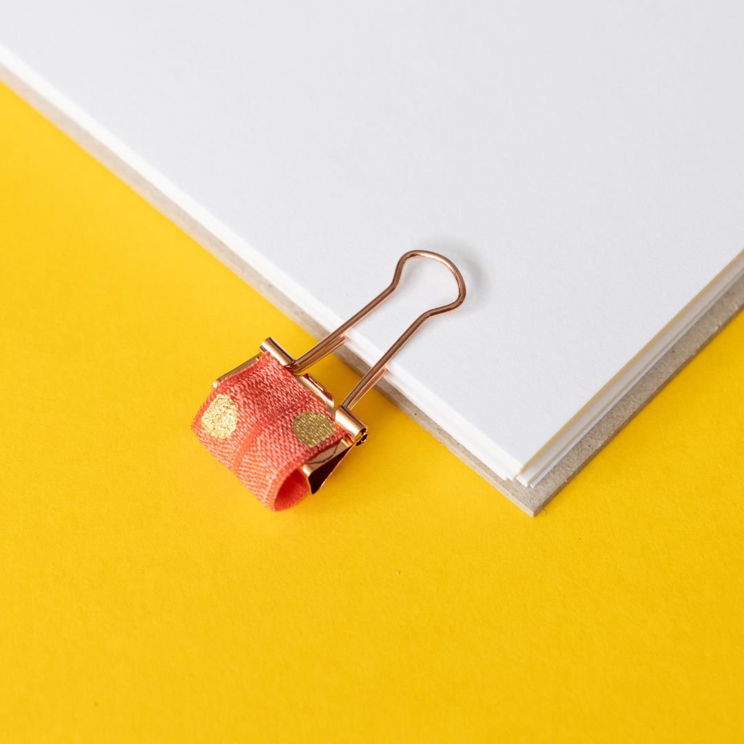Rose gold binder clip with coral and gold elastic on a yellow background