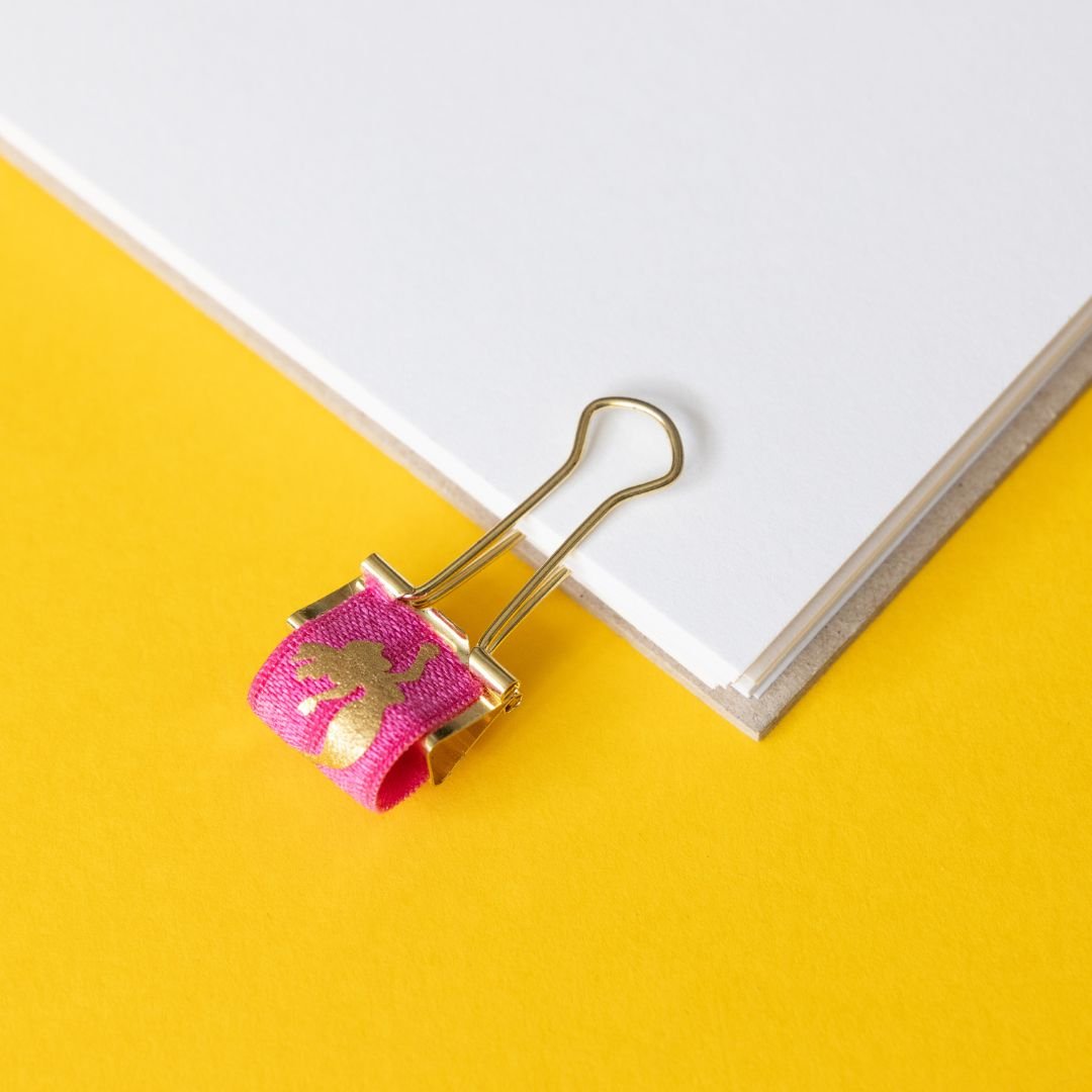 Gold binder clip with pink mermaid elastic on a yellow and white background